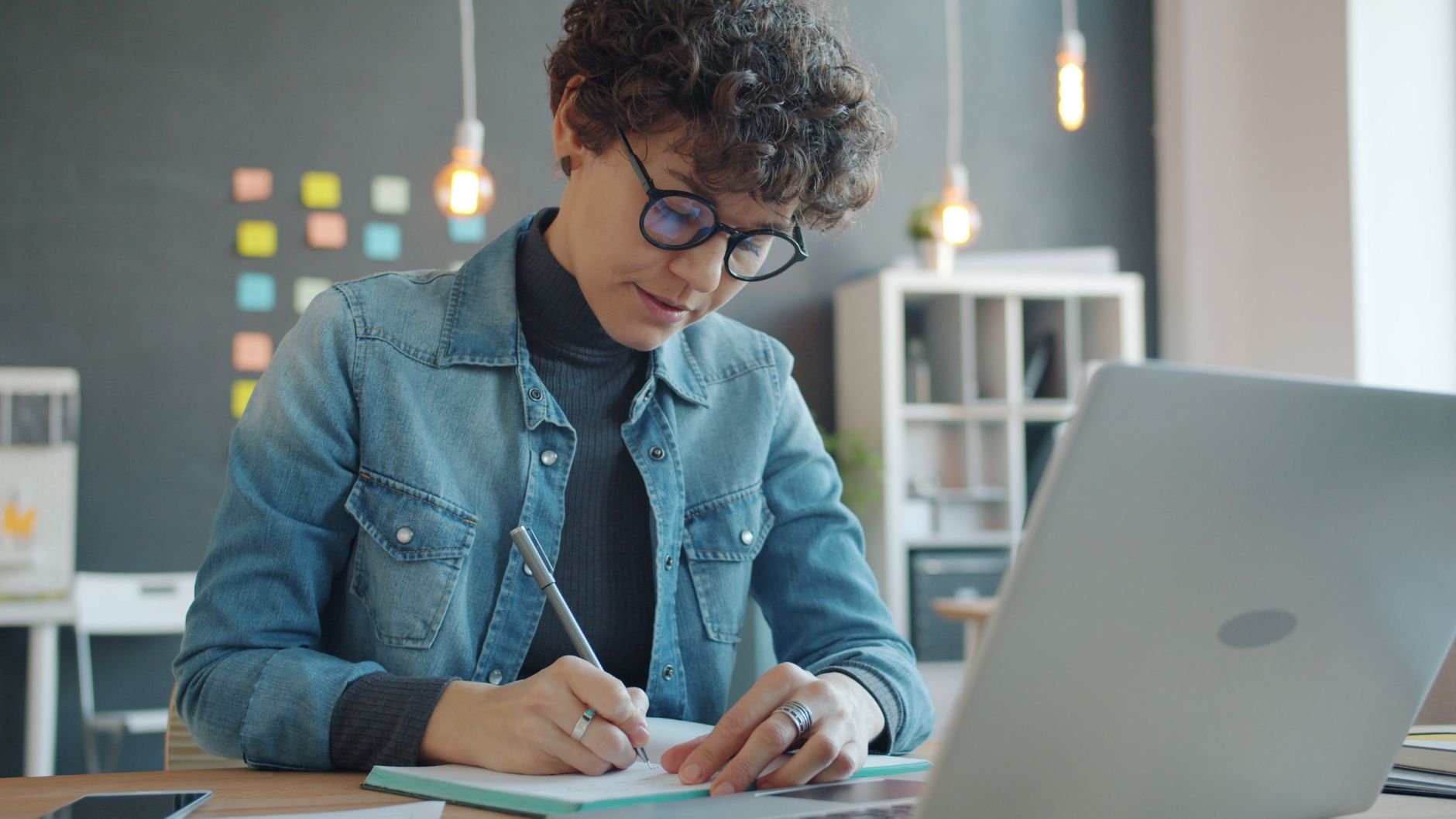Woman writing notes at a desk with a laptop in a modern office setting.