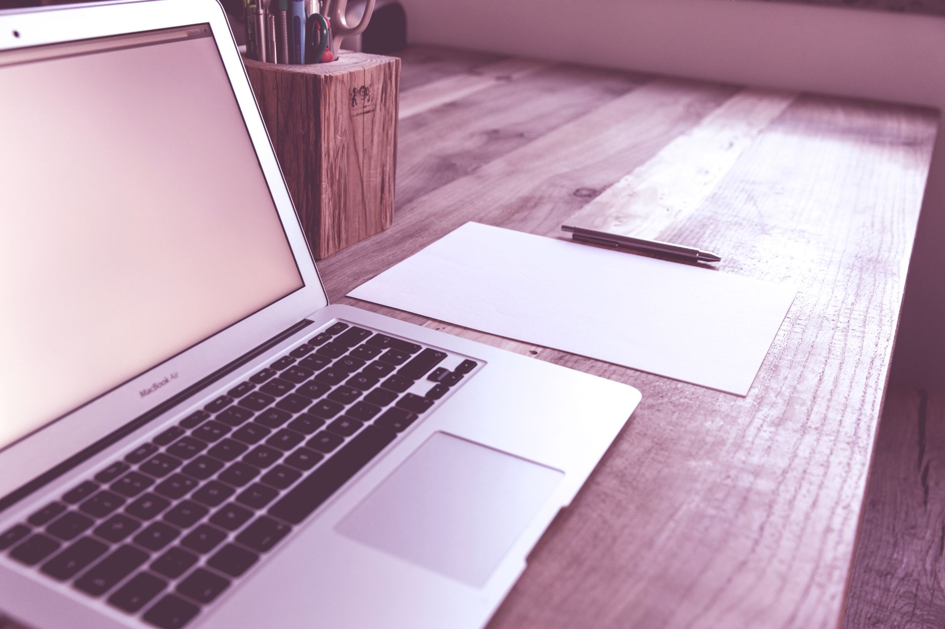 An open laptop on a wooden desk beside a blank sheet of paper with a pen resting on it, and a cup of pens in the background.
