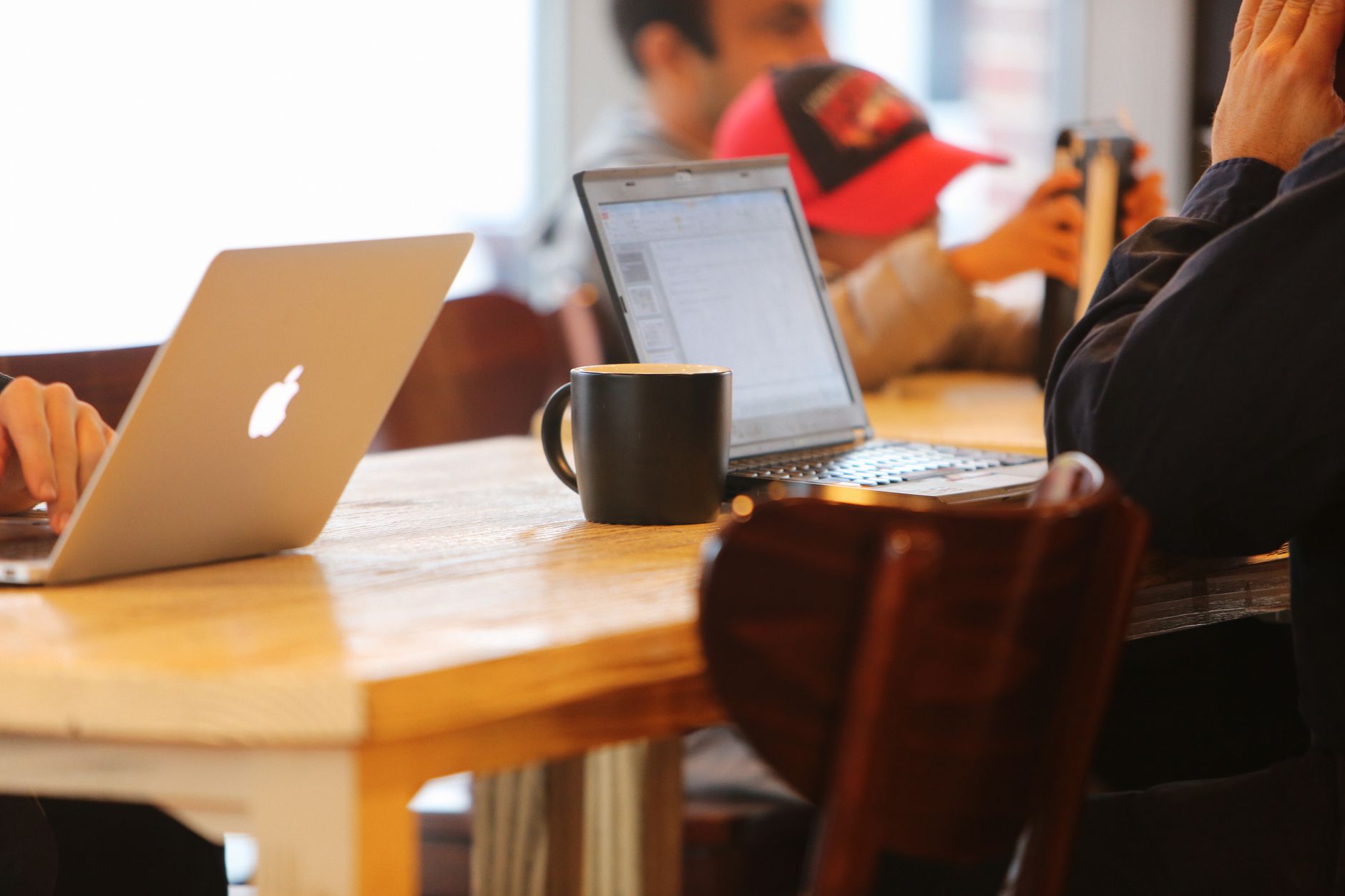 Two open laptops and a black coffee mug on a cafe table, with people seated at nearby tables in the blurred background.