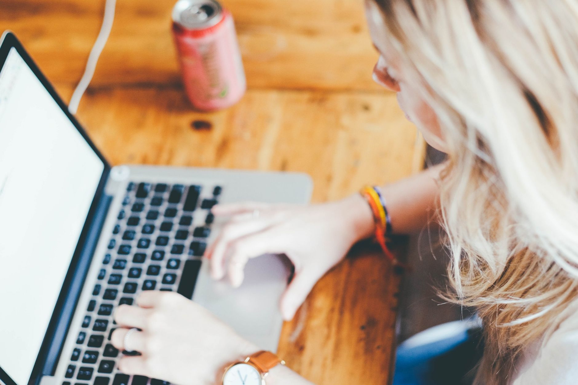 Woman working remotely on a laptop, typing at a wooden desk with a beverage can.