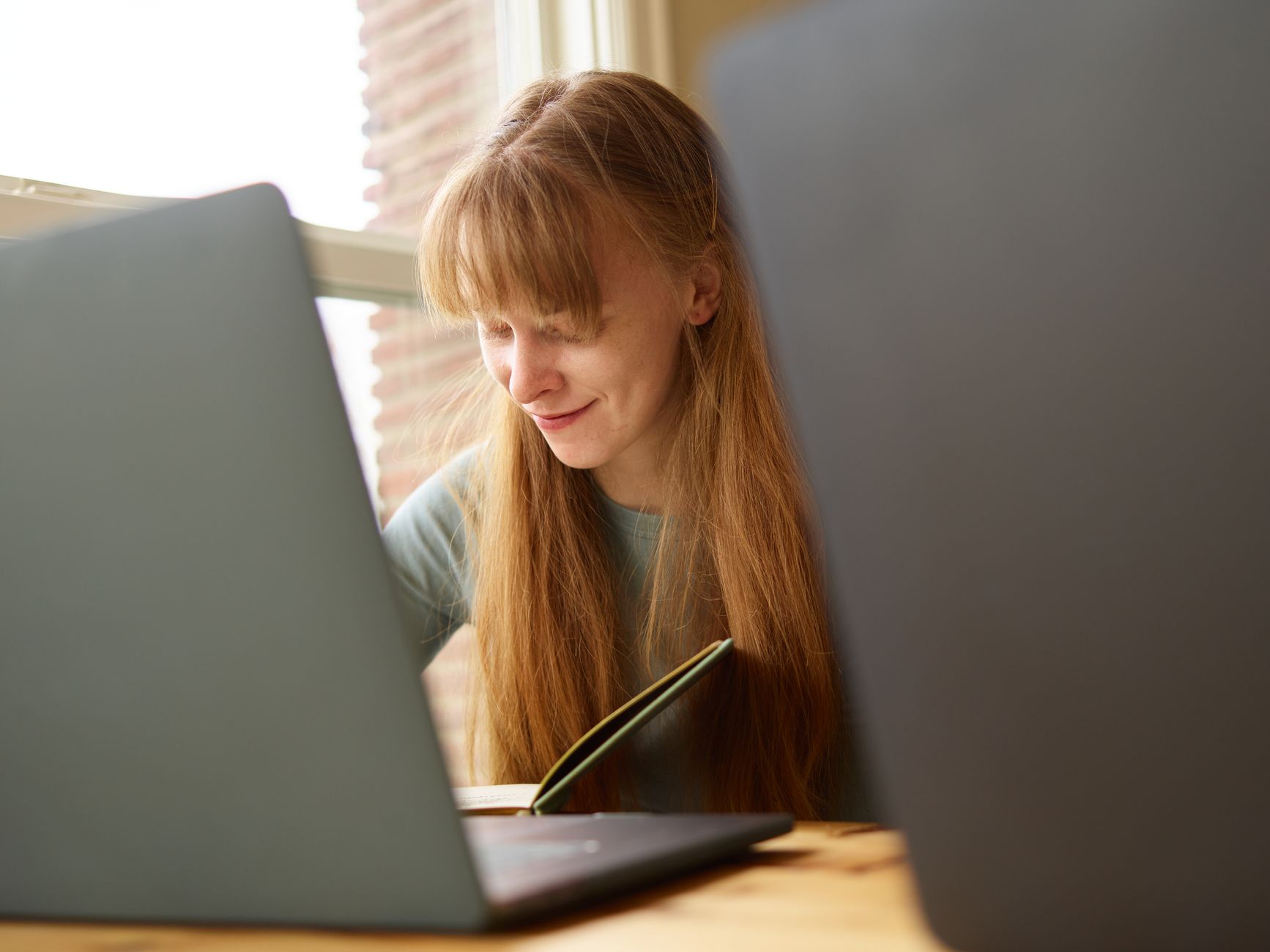 Woman smiling while working on a laptop at a wooden table beside a window.