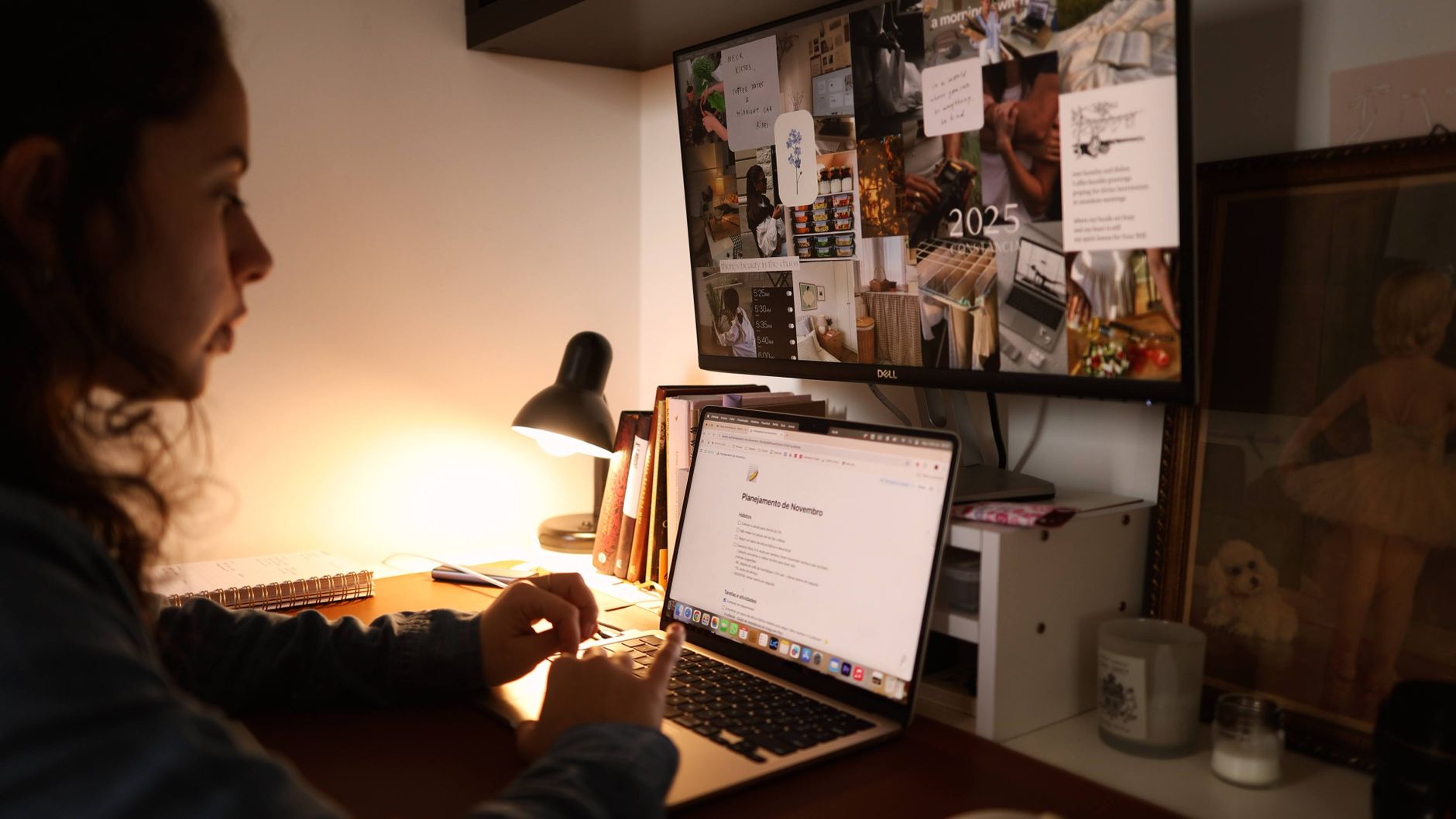 A woman working at a home office desk with a laptop and monitor in warm light.