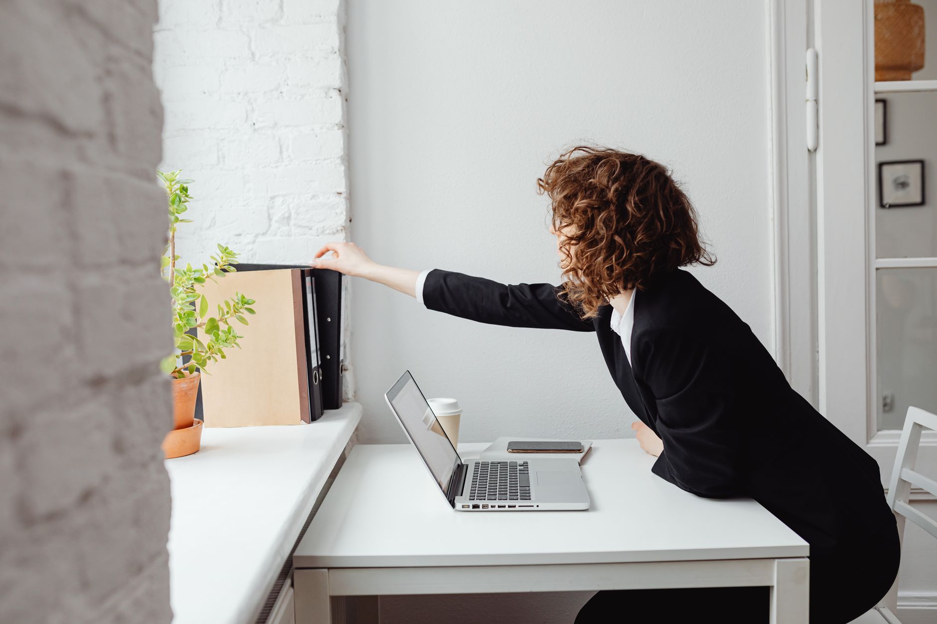 Businesswoman reaching for folders on a shelf while working on a laptop in a bright office.