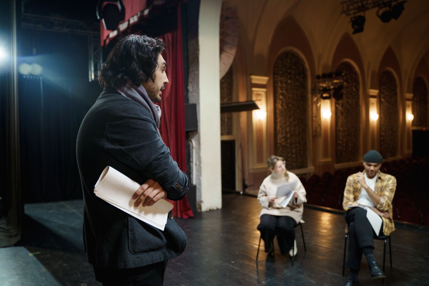 A director with a rolled script watches two actors rehearse on stage, the empty gilded auditorium behind them.