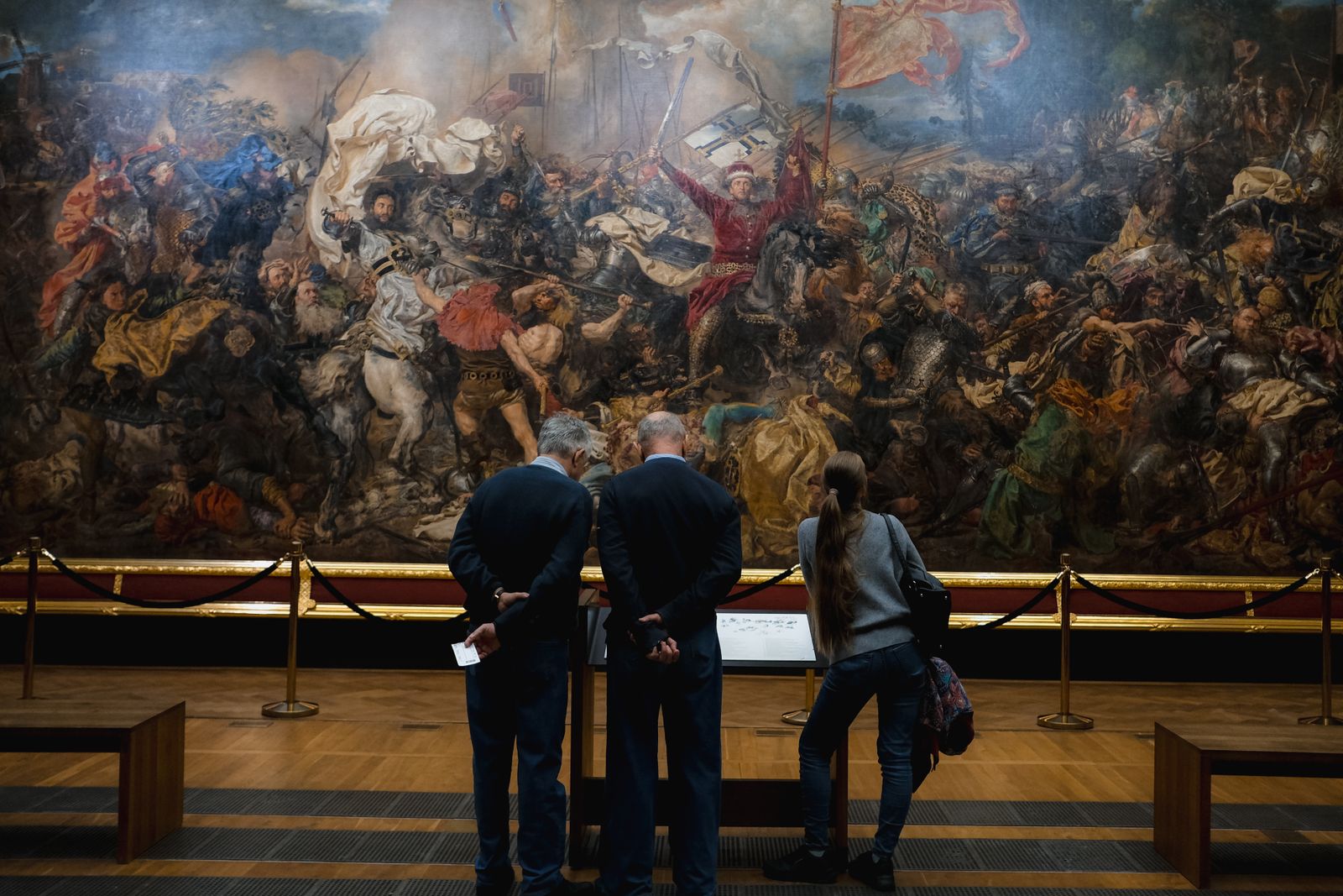 Three visitors at the rope barrier in front of an enormous historical battle painting in a warm, dark museum hall.