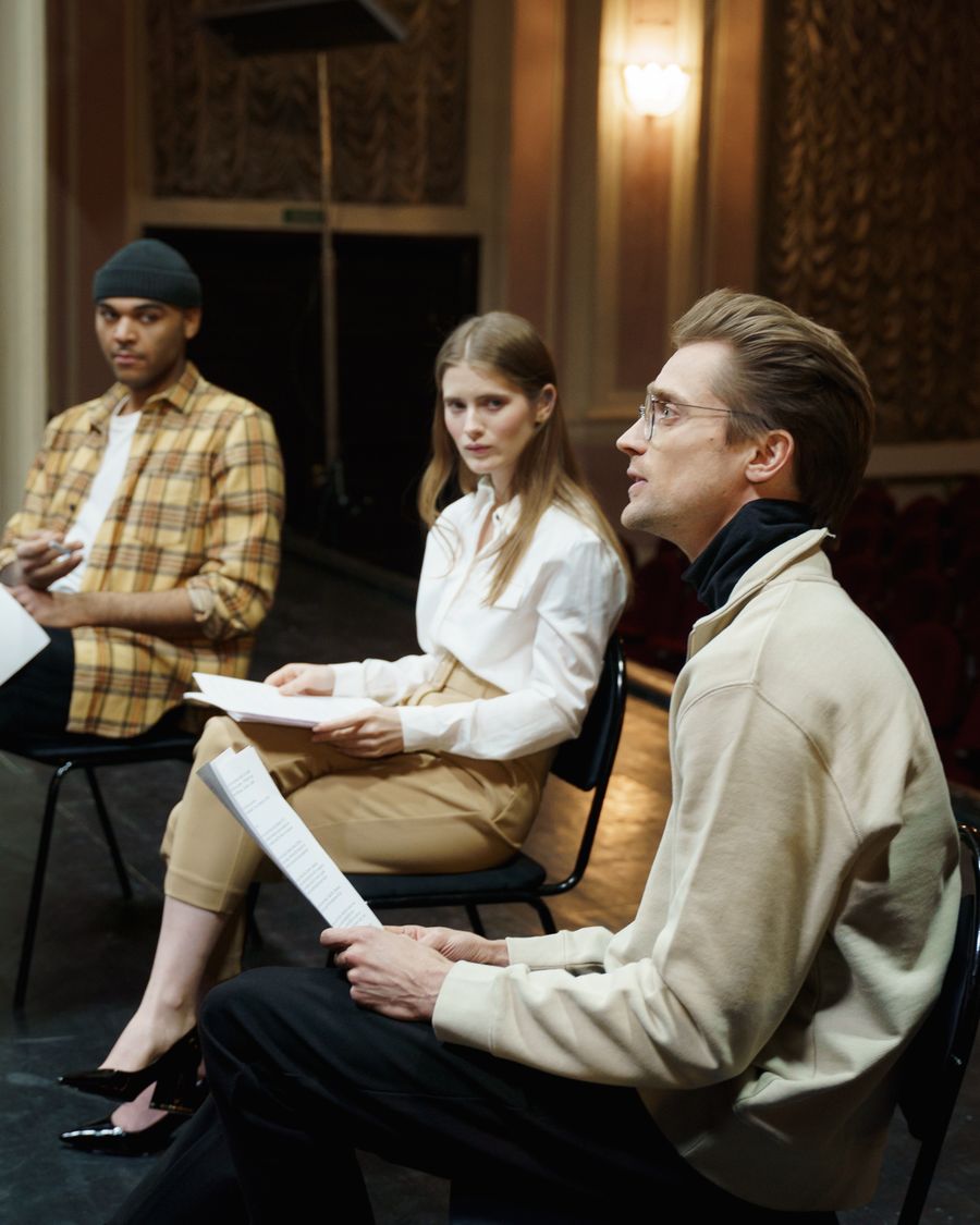 Three actors seated with scripts in their hands during a read-through in a warm, empty theater.