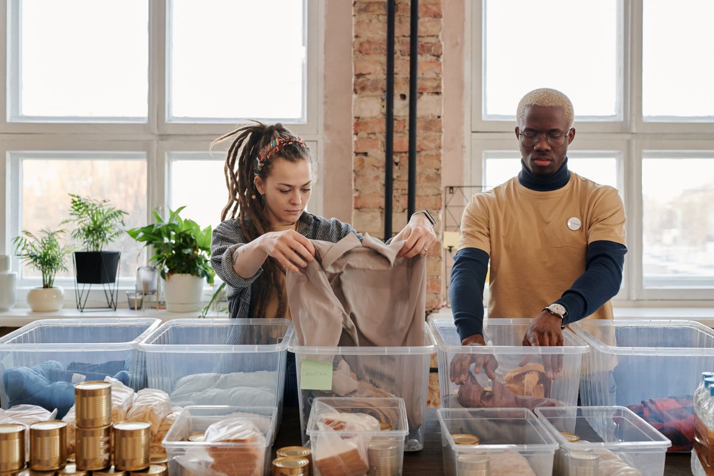 Two volunteers at a table of clear storage boxes, sorting donated clothing and tinned food in front of tall bright windows.