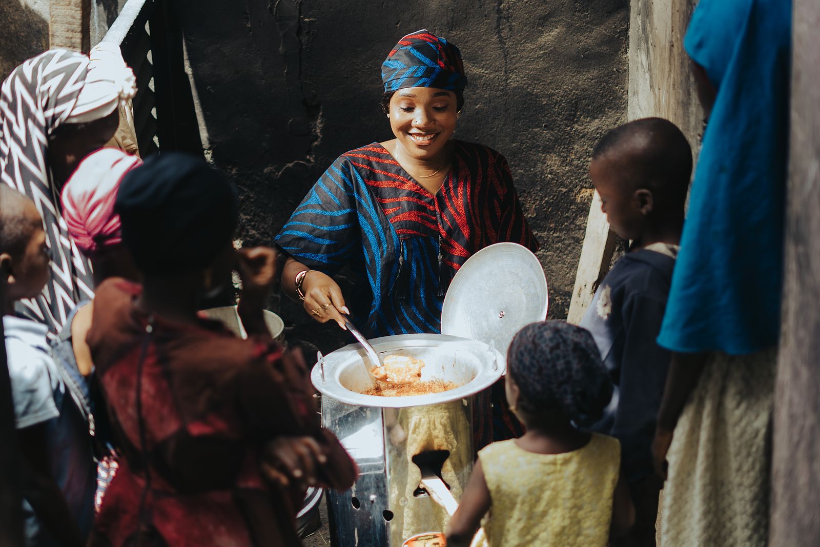 A woman ladling food from a large pot into bowls for a queue of waiting children in an open yard.