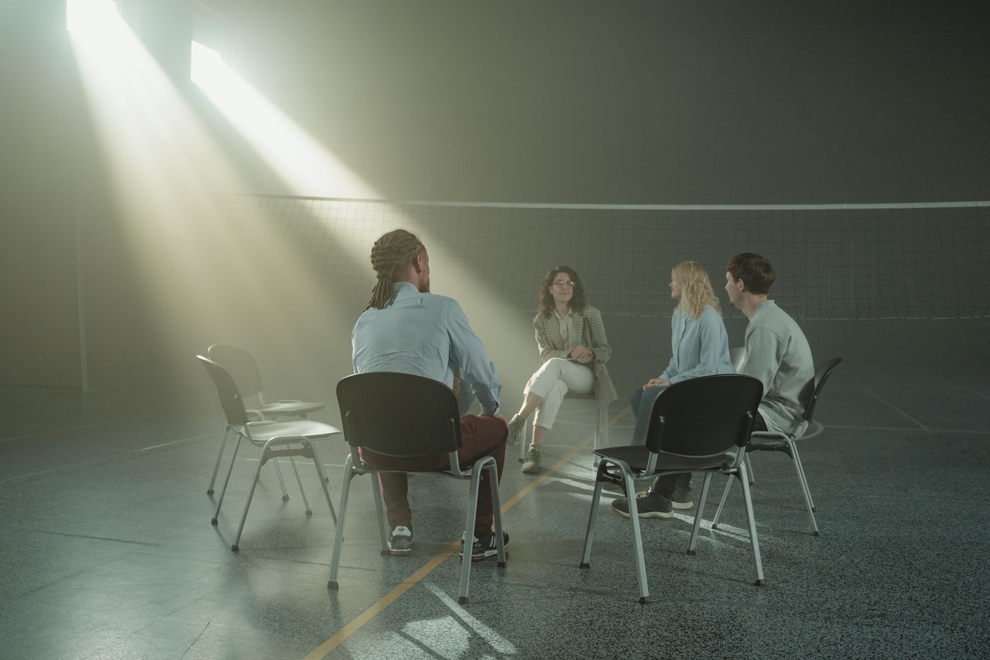 Four people on chairs in a loose circle on a sports-hall floor, sunlight falling in through high windows.