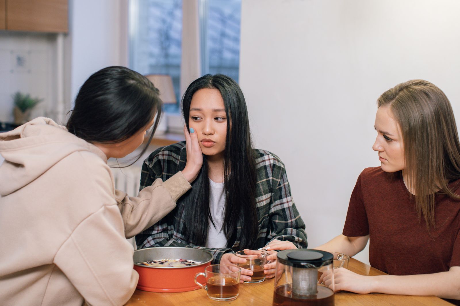 Three people at a kitchen table, two of them comforting a distressed friend over tea.