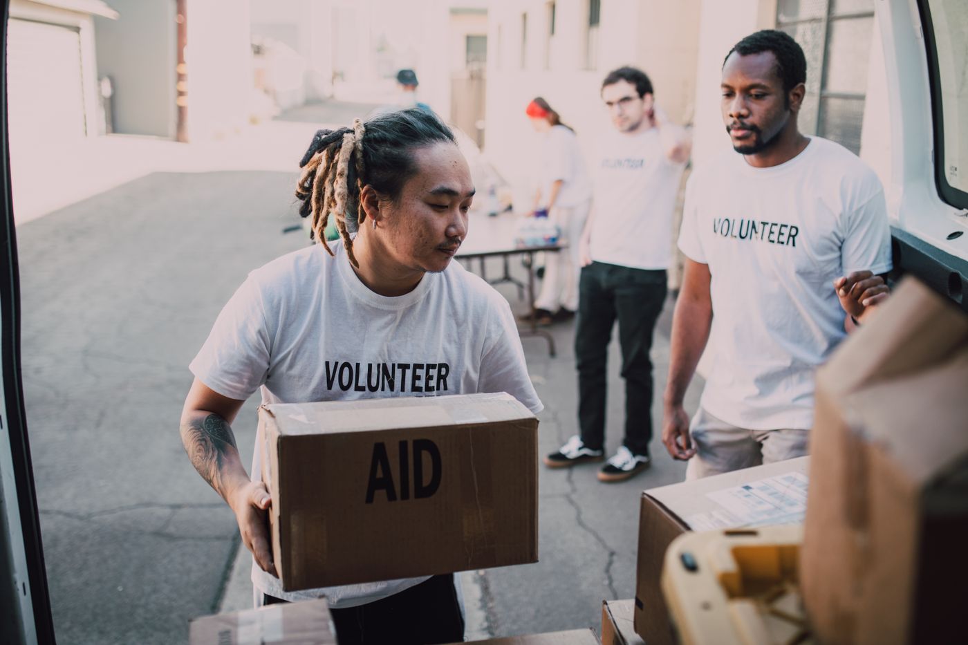 Volunteers in white shirts unloading cardboard boxes marked AID from the back of a van in a sunlit yard.